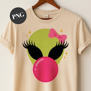 May include: A beige t-shirt with a cartoon alien face. The alien has a green head, black eyelashes, and a pink bubblegum bubble. A pink bow sits atop the alien's head, with yellow star accents.