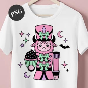 May include: White t-shirt featuring a pink and black cartoon nutcracker holding a cupcake. The nutcracker has a pink hat and uniform with mint green bows. Stars, moons, and a bat surround the nutcracker.