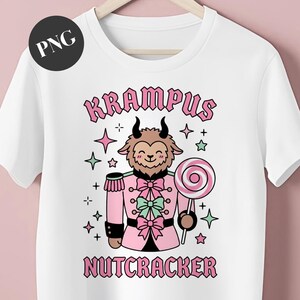 Könnte beinhalten: Weißes T-Shirt mit einem Cartoon-Krampus-Nussknacker-Design. Die Illustration zeigt eine lächelnde ziegenartige Figur in rosa Uniform, die einen Lolli hält, umgeben von Sternen. Das Wort "Krampus" steht über der Figur und "Nussknacker" darunter.