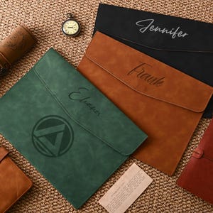 May include: A selection of personalised leather document holders in black, brown, and green. Each holder has a name, such as "Jennifer", "Frank", and "Eleanor", in a stylish script. A vintage pocket watch and a rolled-up map are also visible.