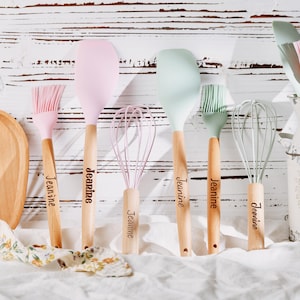 May include: A collection of pastel-colored kitchen utensils with wooden handles, including a spatula, whisk, and brush. Each handle is engraved with the name "Jeanne". A wooden cutting board and a decorative container are also present.