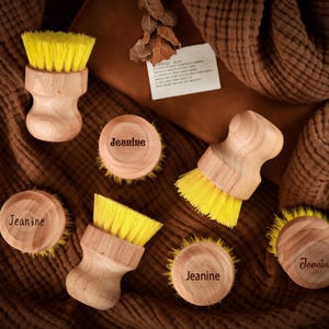 May include: Wooden dish brushes with yellow bristles and a natural wood finish are displayed. Several round wooden soap dishes are also shown, each engraved with the name "Jeanine". The items are arranged on a brown, textured surface.