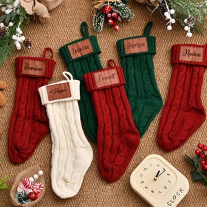 以下が含まれることがあります： 赤、緑、クリーム色のケーブル編みのクリスマスストッキング6つ。各ストッキングには、Jeanine、Puper、Emmett、Thorsten、Radeneという名前の革製のタグが付いています。時計は12時05分を示しています。お祝いの装飾がストッキングを囲んでいます。