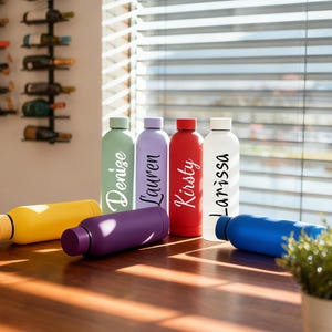 May include: A collection of colorful, personalized water bottles in various shades, including yellow, purple, green, red, white, and blue. Each bottle has a name printed on it in a stylish script font. The bottles are displayed on a wooden table.