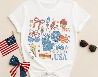 May include: White t-shirt featuring a patriotic design with the Statue of Liberty, fireworks, and the text "USA" and "1776". The shirt also includes illustrations of a cupcake, ice cream cone, and a hot dog. An American flag and bow are also present.