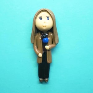Peut inclure: Petite figurine représentant une personne aux longs cheveux blonds, vêtue d'une veste marron, d'un pantalon noir et de chaussures marron. La figurine tient une sphère bleue. Le fond est de couleur turquoise.