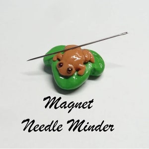 May include: A green frog sitting on a green leaf with a needle stuck in it. The frog and leaf are magnetic and are used as a needle minder for sewing.