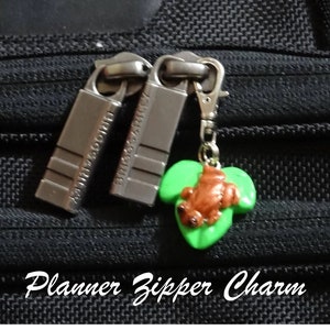 May include: A green leaf charm with a brown frog on it, attached to a silver zipper pull. The charm is labeled "Planner Zipper Charm".