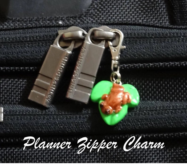 May include: A green leaf charm with a brown frog on it, attached to a silver zipper pull. The charm is labeled "Planner Zipper Charm".