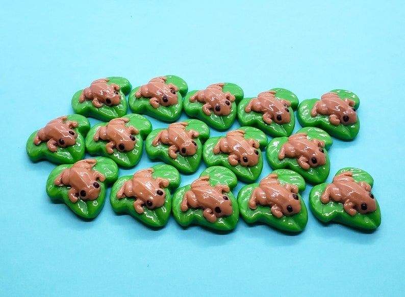 May include: A collection of 15 brown frog figurines on green lily pad bases. The frogs are all in a similar pose, with their legs outstretched and their heads tilted up.