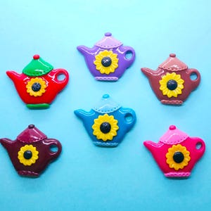 May include: Six colourful teapot-shaped charms with a sunflower design. The charms are made of plastic and have a glossy finish. The colours of the charms are purple, red, brown, blue, pink, and brown.