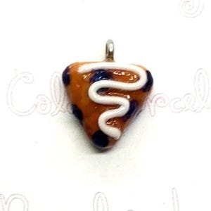 May include: A handmade pendant in the shape of a slice of pizza. The pendant is orange with dark blue spots and a white, squiggly line across the top. It has a silver-colored loop at the top for attaching to a chain.