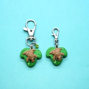 May include: Two silver keychains with green leaf charms featuring brown frog figurines. The larger keychain has a green rhinestone.