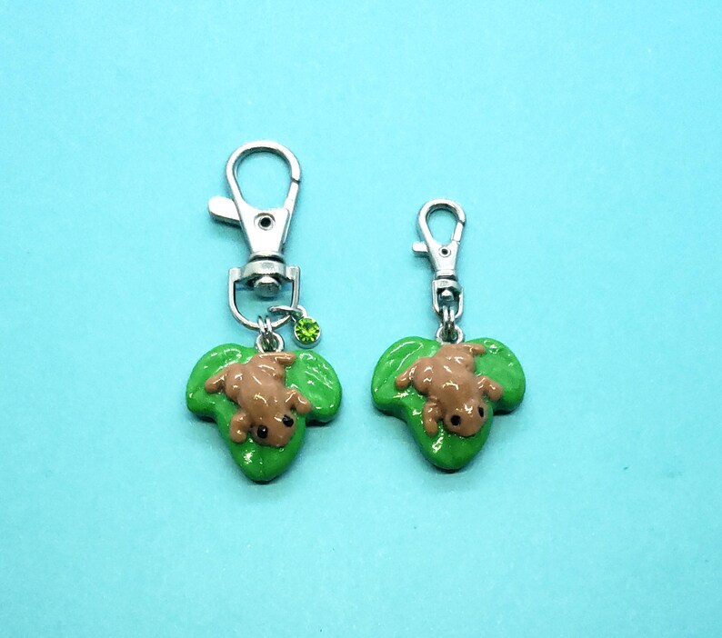 May include: Two silver keychains with green leaf charms featuring brown frog figurines. The larger keychain has a green rhinestone.