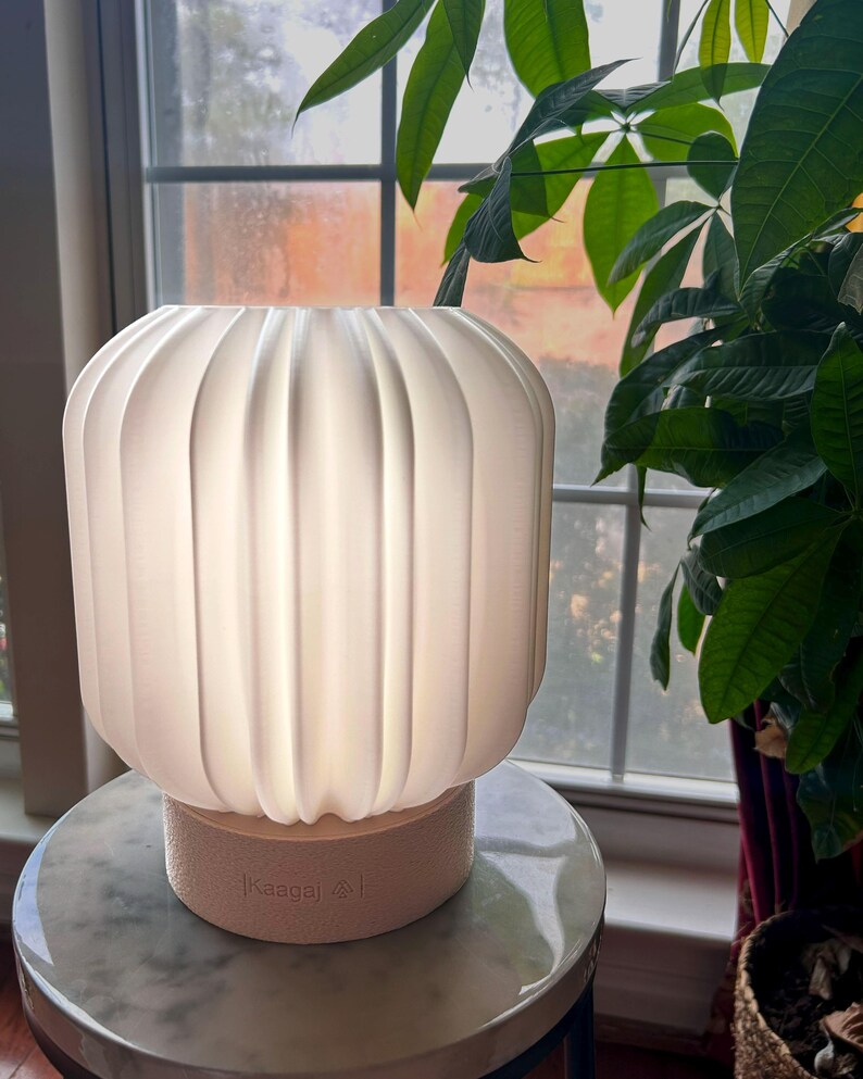 May include: A white, ribbed, cylindrical lamp with a soft glow. The lamp sits on a light beige base with the text "|Kaaga| &and; |" printed on it. The lamp is on a marble-topped table, with green plants in the background.