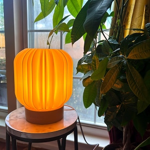 May include: A glowing orange table lamp with a ribbed shade sits on a small round table. The lamp's base is a light brown color. The lamp is plugged into an electrical outlet. The background includes a window and lush green plants.
