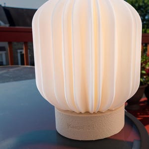 May include: A glowing, off-white lamp with a ribbed, rounded shade. The lamp sits on a beige base with the text "Kaagaj" printed on it. The lamp is on a glass table outdoors.
