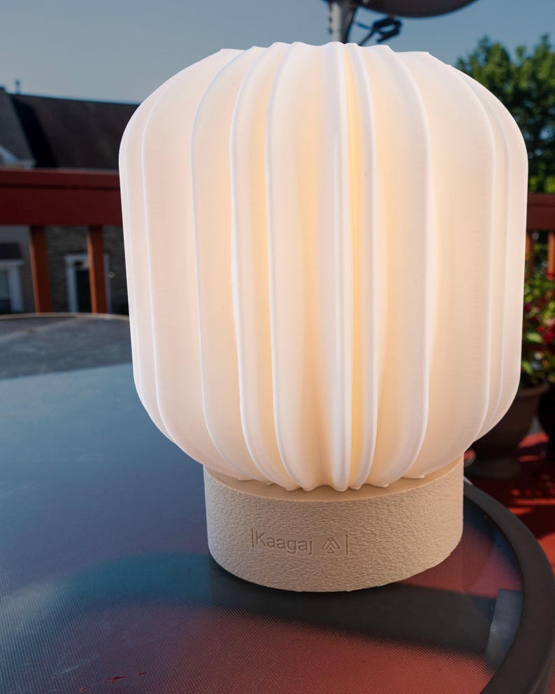 May include: A glowing, off-white lamp with a ribbed, rounded shade. The lamp sits on a beige base with the text "Kaagaj" printed on it. The lamp is on a glass table outdoors.