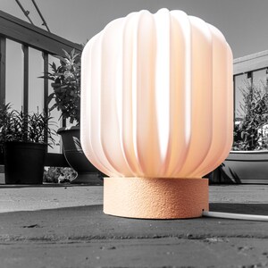 May include: A peach-colored, ribbed table lamp with a textured base. The lamp has a rounded, fluted shade and is lit from within, casting a warm glow. The lamp is on a wooden deck, with plants in the background.