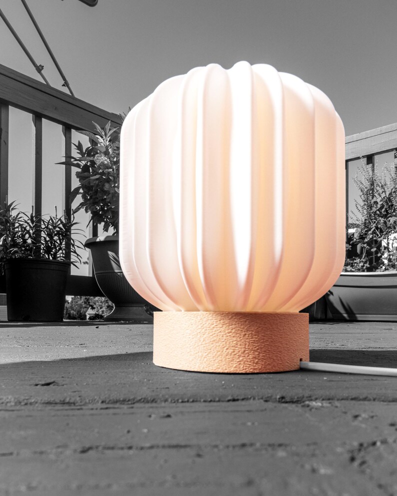 May include: A peach-colored, ribbed table lamp with a textured base. The lamp has a rounded, fluted shade and is lit from within, casting a warm glow. The lamp is on a wooden deck, with plants in the background.