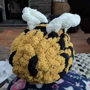 May include: A round, plush, crocheted bee toy featuring a textured yellow and black striped body. The bee has two prominent, fluffy white wings attached to the top, crafted from soft, fuzzy yarn that creates a distinct, tactile appearance.