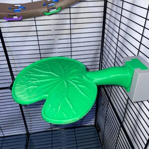 May include: A bright green lily pad platform for a small animal cage, featuring a textured leaf surface and a sturdy stem attached to a gray rectangular mounting block, positioned against a black wire grid wall inside an enclosure.