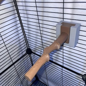 May include: A brown, textured, branch-shaped perch attached to a light gray, square mounting bracket. The perch is secured to a black wire cage frame. The branch features a realistic, knobby surface texture and extends outward from the mount.