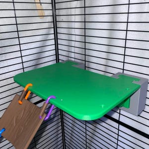May include: A green rectangular platform with rounded corners attached to a black wire cage wall by two gray mounting blocks. A brown textured panel hangs from the front edge of the platform, secured by one orange and one purple plastic link.