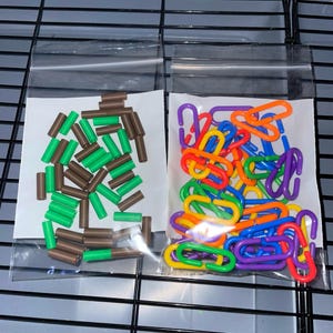 以下が含まれることがあります： クラフト用品が入った2つの透明なビニール袋。左の袋には茶色と緑色の円筒形のプラスチックビーズが入っています。右の袋には、明るいオレンジ、黄色、緑、青、紫の連結可能なプラスチック製のチェーンリンクが入っています。