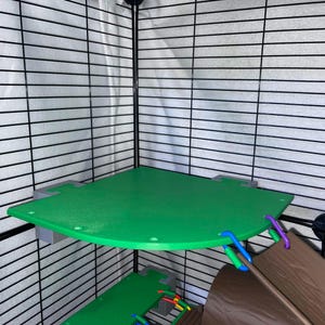 May include: A green corner platform for a small animal cage, featuring gray mounting brackets and a curved front edge. The platform includes small circular holes along the perimeter and is attached to a black wire cage wall with colorful plastic links.
