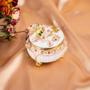 May include: A white, round trinket box with a floral design. The box features pink and gold accents, with a butterfly on top. The box is adorned with pink flowers and rhinestones. The box sits on three gold feet.
