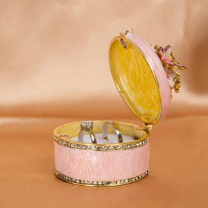 Puede incluir: Una caja de anillos rosa con un borde dorado y un dise&ntilde;o de mariposa en la tapa. La caja est&aacute; abierta y hay dos anillos dentro.