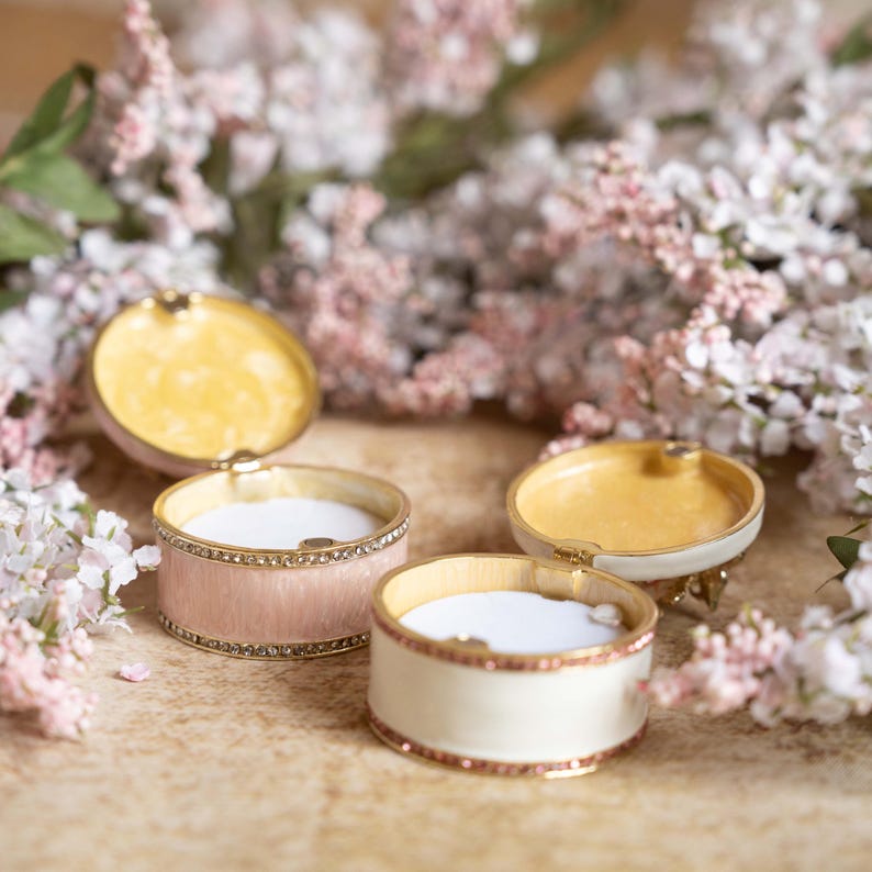 Puede incluir: Tres peque&ntilde;as cajas redondas decorativas con tapas. Las cajas son de metal y tienen un acabado de esmalte blanco, rosa y crema. Las tapas est&aacute;n abiertas y revelan un interior amarillo. Las cajas est&aacute;n decoradas con un borde dorado y diamantes de imitaci&oacute;n.