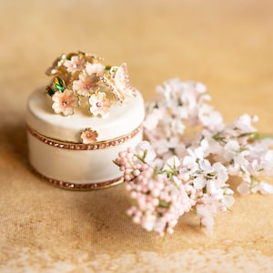 Puede incluir: Una caja de porcelana blanca para guardar peque&ntilde;os objetos con un dise&ntilde;o floral que presenta flores rosas y blancas y mariposas. La caja est&aacute; decorada con pedrer&iacute;a y tiene una tapa con bisagras.