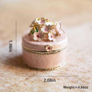 Puede incluir: Una peque&ntilde;a caja de joyer&iacute;a rosa con un dise&ntilde;o floral. La tapa est&aacute; adornada con un grupo de flores rosas con hojas verdes y detalles dorados. La caja mide aproximadamente 5 cm de di&aacute;metro y 5 cm de alto. Pesa aproximadamente 138 gramos.