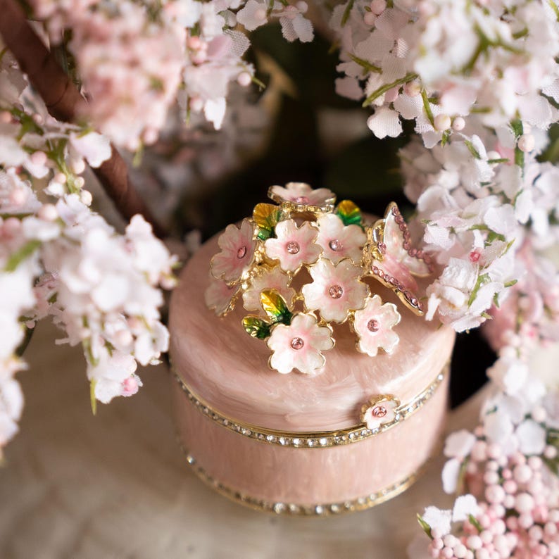 Puede incluir: Una caja de joyas rosa con un dise&ntilde;o floral. La tapa est&aacute; adornada con un grupo de flores rosas y una mariposa. La caja est&aacute; decorada con una fila de diamantes de imitaci&oacute;n alrededor de la base.