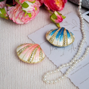 May include: Two decorative seashell-shaped trinket boxes with gold-tone metal edges. One shell features white and pink enamel with embedded clear rhinestones, while the other features white and blue enamel with embedded clear rhinestones.