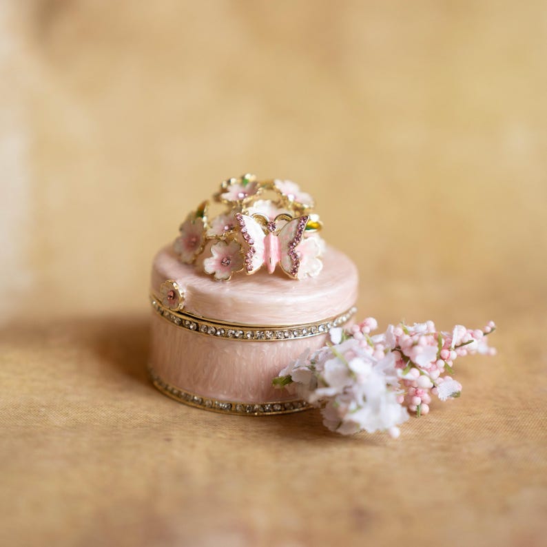Puede incluir: Una caja de joyas rosa con un dise&ntilde;o floral y una mariposa en la parte superior. La caja est&aacute; decorada con diamantes de imitaci&oacute;n y tiene un borde dorado.