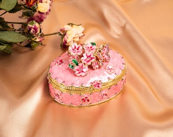 May include: Pink oval jewelry box with floral and butterfly embellishments. The box features a gold-colored trim and is adorned with pink flowers and a butterfly on the lid. The box is set against a satin background.