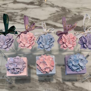 May include: Six small decorative boxes with hydrangea flowers on top. The boxes are in shades of pink, purple, and blue. The flowers are made of fabric and have a realistic appearance. The boxes are arranged in two rows of three. The boxes are labeled with the words "Love is Eternal" in gold lettering.