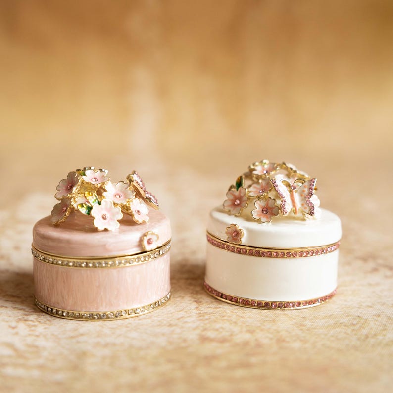 Puede incluir: Dos peque&ntilde;as cajas decorativas con dise&ntilde;os florales y de mariposas. La caja de la izquierda es rosa con un borde dorado y la caja de la derecha es blanca con un borde dorado.