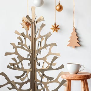 K&ouml;nnte beinhalten: Ein dekorativer Weihnachtsbaum aus Holz mit offenem Design steht vor einer wei&szlig;en Wand. H&auml;ngende Dekorationen umfassen eine wei&szlig;e Kugel, eine goldene Kugel, einen goldenen Schneeflocken-Anh&auml;nger und einen Holzbaum. Eine wei&szlig;e Tasse steht auf einem kleinen Holzschemel.