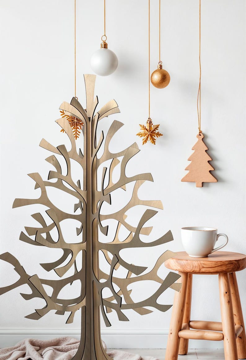 K&ouml;nnte beinhalten: Ein dekorativer Weihnachtsbaum aus Holz mit offenem Design steht vor einer wei&szlig;en Wand. H&auml;ngende Dekorationen umfassen eine wei&szlig;e Kugel, eine goldene Kugel, einen goldenen Schneeflocken-Anh&auml;nger und einen Holzbaum. Eine wei&szlig;e Tasse steht auf einem kleinen Holzschemel.