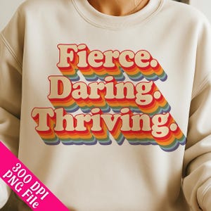 Op de afbeelding: Een wit sweatshirt met een regenboogkleurige afbeelding met de tekst "Fierce. Daring. Thriving."