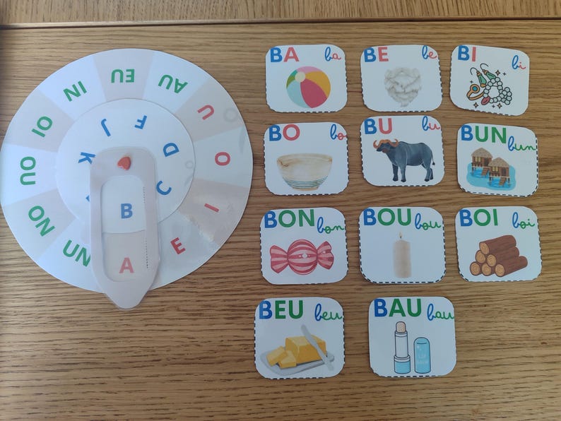 May include: A colorful learning wheel with letters and pictures. The wheel has a spinning dial with letters A, B, C, D, E, F, J, K, L, N, O, U, and the words 'IN', 'EU', 'AU', 'OU', 'ON', 'UN', 'OI', 'OU'. The cards have pictures of a beach ball, a heart, a necklace, a bowl, a buffalo, a hut, a candy, a candle, logs, a knife and butter, and a lipstick.