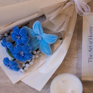 May include: A bouquet of blue velvet flowers with white accents, wrapped in cream-colored paper and tied with a white ribbon. A scented candle and a book titled "The Art of Home" are also present.