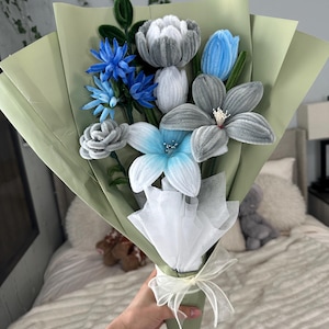 May include: A bouquet of handmade felt flowers in shades of blue, gray, and white, wrapped in light green paper. The bouquet includes various flower types, such as tulips and lilies, and is tied with a white ribbon. The flowers are held in a cone shape.