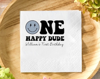 Könnte beinhalten: Weiße quadratische Papierserviette mit dem Text "ONE HAPPY DUDE William's First Birthday" in Schwarz und einem Smiley mit Blitzen. Die Serviette liegt auf einem Holztablett mit Grünzeug.