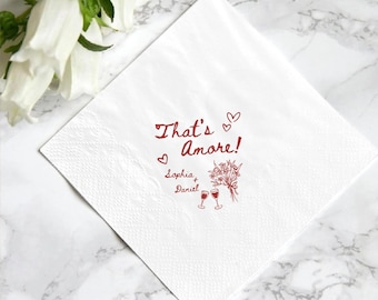 May include: White paper napkin with the text "That's Amore!" in red script, along with hearts, a bouquet of flowers, and two wine glasses. The names "Sophia + Daniel" are also printed on the napkin.