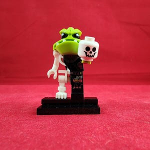 May include: A Lego minifigure featuring a green frog-like head, white arms and legs, and a black torso. The figure holds a white skull head. It stands on a black base against a red background.